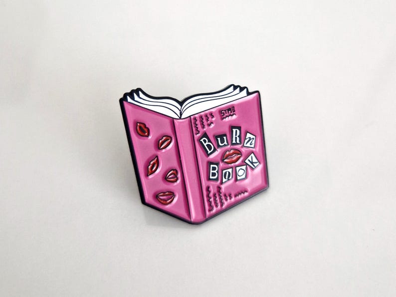 Puede incluir: Pin de esmalte rosa en forma de libro con las palabras "Burn Book" en la portada. El libro tiene labios rojos y corazones en la portada.