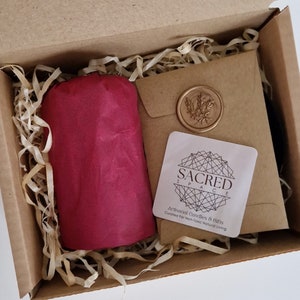 K&ouml;nnte beinhalten: Ein brauner Karton mit einem in rotes Seidenpapier eingewickelten Geschenk darin. Die Schachtel ist mit zerkleinertem Papier gef&uuml;llt. Ein kleines wei&szlig;es quadratisches Etikett mit dem Text "SACRED SPACE" und den Worten "Artisanal Candles & Gifts Curated for Non-Toxic Natural Living" ist sichtbar.