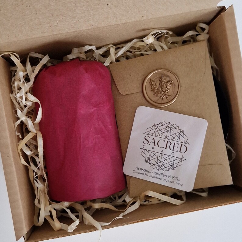 K&ouml;nnte beinhalten: Ein brauner Karton mit einem in rotes Seidenpapier eingewickelten Geschenk darin. Die Schachtel ist mit zerkleinertem Papier gef&uuml;llt. Ein kleines wei&szlig;es quadratisches Etikett mit dem Text "SACRED SPACE" und den Worten "Artisanal Candles & Gifts Curated for Non-Toxic Natural Living" ist sichtbar.
