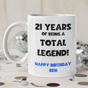 K&ouml;nnte beinhalten: Eine wei&szlig;e Keramiktasse mit schwarzem Text, der "21 Years of Being a Total Legend! Happy Birthday Ben" lautet. Die Tasse steht auf einer wei&szlig;en Holzoberfl&auml;che mit silbernem Konfetti und Discokugeln.