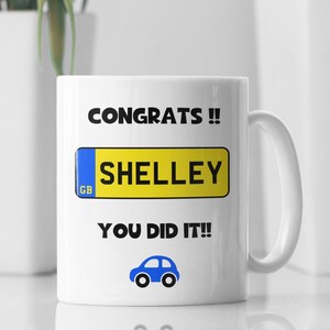 May include: White ceramic mug with a blue and yellow car number plate design. The number plate says "CONGRATS!! SHELLEY YOU DID IT!!" with a small blue car icon below the text.