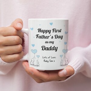 Puede incluir: Taza de cer&aacute;mica blanca con una ilustraci&oacute;n azul y blanca de elefantes sosteniendo globos en forma de coraz&oacute;n azul. La taza tiene el texto "Happy First Father's Day as my Daddy Lots of Love, Baby Tom x".