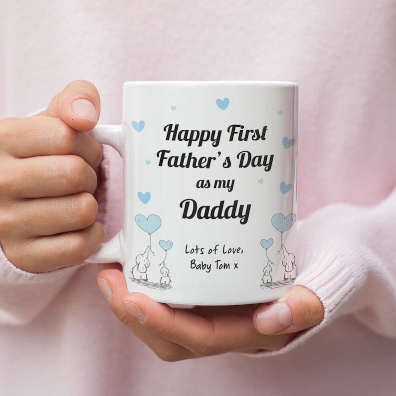 Puede incluir: Taza de cer&aacute;mica blanca con una ilustraci&oacute;n azul y blanca de elefantes sosteniendo globos en forma de coraz&oacute;n azul. La taza tiene el texto "Happy First Father's Day as my Daddy Lots of Love, Baby Tom x".