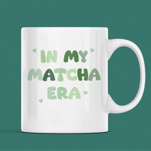 Może przedstawiać: Biały kubek ceramiczny z napisem 'In My Matcha Era' w zielono-białych literach. Tekst jest w zabawnym, zaokrąglonym kroju pisma.