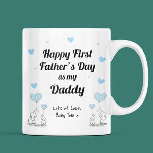 Puede incluir: Taza de cer&aacute;mica blanca con un dise&ntilde;o azul y negro. La taza dice "Happy First Father's Day as my Daddy" con dos corazones azules y dos elefantes que sostienen globos en forma de coraz&oacute;n azules. El texto "Lots of Love, Baby Tom x" est&aacute; en la parte inferior de la taza.