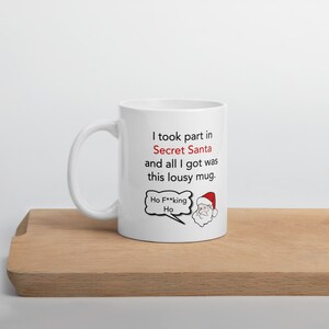 Op de afbeelding: Witte keramische mok met een handvat. De mok heeft de tekst "I took part in Secret Santa and all I got was this lousy mug." Een cartoon kerstman met een tekstballon met "Ho F**king Ho" staat ook op de mok.