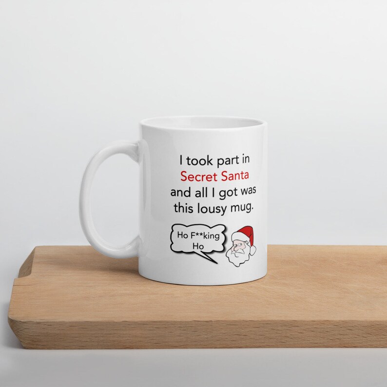 Op de afbeelding: Witte keramische mok met een handvat. De mok heeft de tekst "I took part in Secret Santa and all I got was this lousy mug." Een cartoon kerstman met een tekstballon met "Ho F**king Ho" staat ook op de mok.