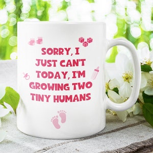 K&ouml;nnte beinhalten: Eine wei&szlig;e Keramiktasse mit einem rosafarbenen Design, auf dem "Sorry, ich kann heute nicht, ich z&uuml;chte zwei kleine Menschen" steht. Die Tasse ist von wei&szlig;en Bl&uuml;ten und gr&uuml;nen Bl&auml;ttern umgeben.