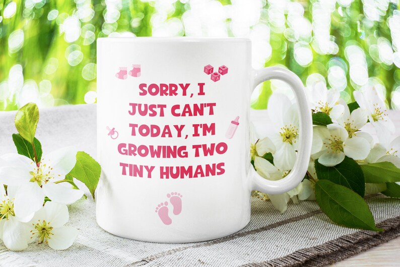 K&ouml;nnte beinhalten: Eine wei&szlig;e Keramiktasse mit einem rosafarbenen Design, auf dem "Sorry, ich kann heute nicht, ich z&uuml;chte zwei kleine Menschen" steht. Die Tasse ist von wei&szlig;en Bl&uuml;ten und gr&uuml;nen Bl&auml;ttern umgeben.
