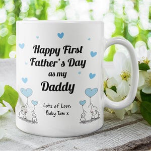 Puede incluir: Taza de cer&aacute;mica blanca con un dise&ntilde;o azul y negro. La taza dice "Happy First Father's Day as my Daddy" con corazones azules y elefantes. El texto "Lots of Love, Baby Tom x" est&aacute; en la parte inferior.