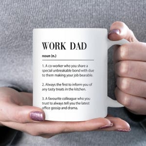 May include: White ceramic mug with the text "WORK DAD" and a definition. The definition includes three points about a co-worker. The mug is held by a person with manicured nails.
