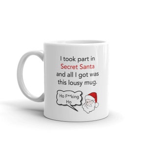 Op de afbeelding: Witte keramische mok met een humoristische boodschap over Secret Santa. De mok heeft de tekst "I took part in Secret Santa and all I got was this lousy mug." Een cartoon kerstman met een tekstballon met "Ho F***ing Ho" staat ook op de mok.