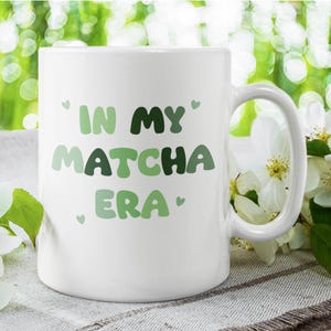 Może przedstawiać: Biały kubek ceramiczny z napisem "In My Matcha Era" w zielono-białych literach. Kubek ma uchwyt i stoi na jasnobrązowej powierzchni.