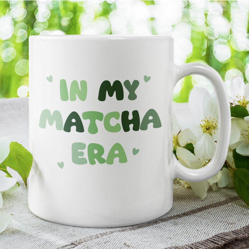 Może przedstawiać: Biały kubek ceramiczny z napisem "In My Matcha Era" w zielono-białych literach. Kubek ma uchwyt i stoi na jasnobrązowej powierzchni.