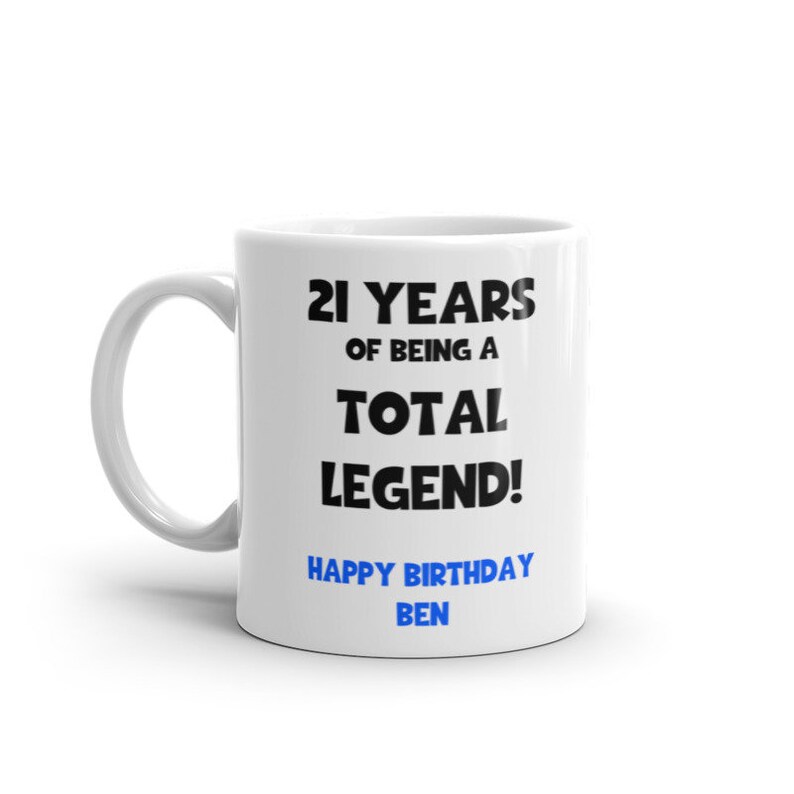 K&ouml;nnte beinhalten: Wei&szlig;e Keramiktasse mit schwarzem Text, der "21 Jahre, ein echter Held! Alles Gute zum Geburtstag Ben" lautet. Der Text "Ben" ist blau.
