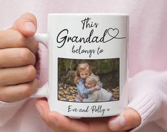 May include: White ceramic mug featuring the text "This Grandad belongs to" above a photograph of two young girls. The names "Eve and Polly" are printed below. The mug is held in a person's hands.