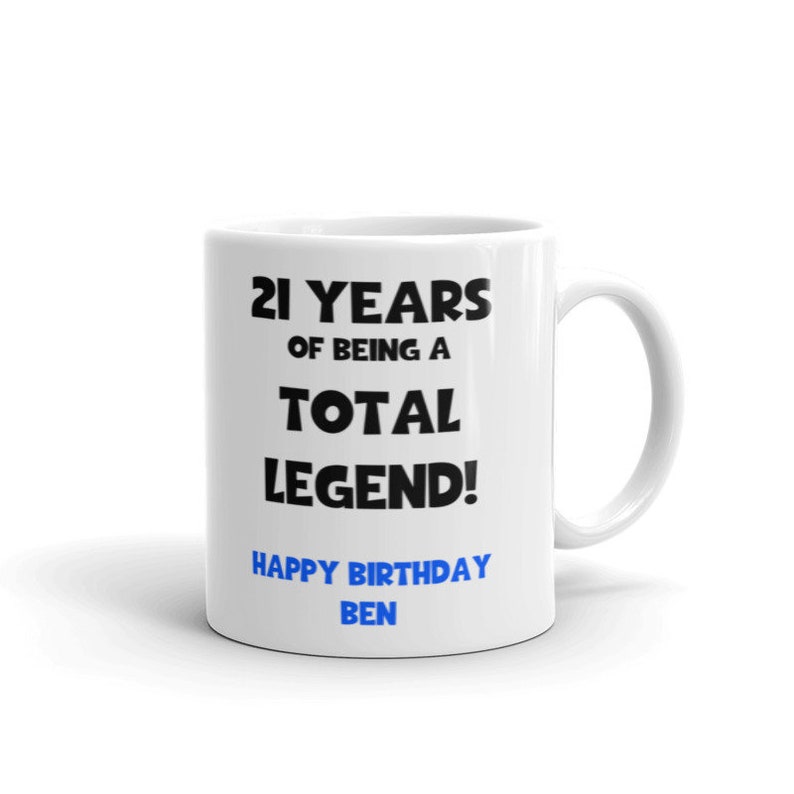 K&ouml;nnte beinhalten: Eine wei&szlig;e Keramiktasse mit schwarzem Text, der "21 Years of Being a Total Legend! Happy Birthday Ben" lautet. Der Text "Happy Birthday" ist blau.