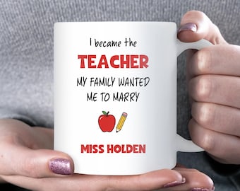 May include: A white ceramic mug featuring the black text I became the TEACHER MY FAMILY WANTED ME TO MARRY, with the word TEACHER in red. Below the text is a red apple and a yellow pencil, followed by the name MISS HOLDEN in red text.
