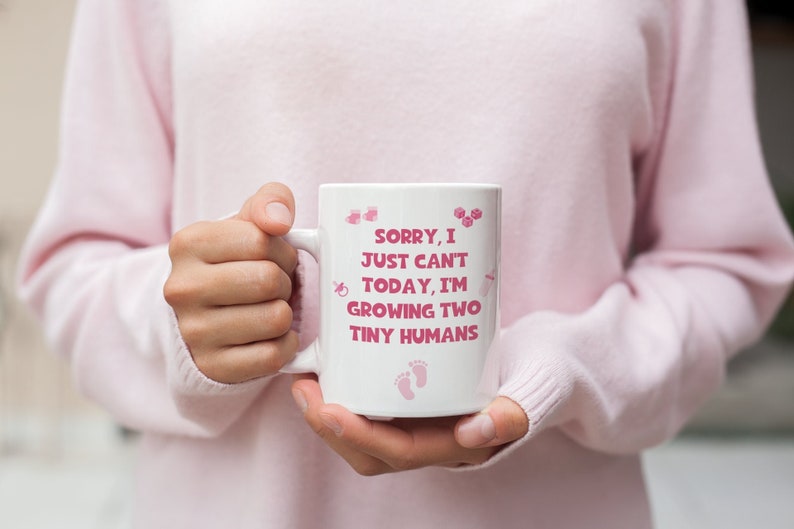 K&ouml;nnte beinhalten: Eine wei&szlig;e Keramiktasse mit einem rosa Henkel und rosa Text, der "Sorry, I just can't today, I'm growing two tiny humans" lautet. Die Tasse ist mit rosa Baby-Symbolen verziert.
