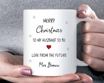 May include: White ceramic mug with the text "MERRY Christmas TO MY HUSBAND TO BE" and "LOVE FROM THE FUTURE Mrs Brown" in black script. A red heart and snowflakes are also featured on the mug.