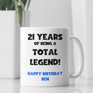 K&ouml;nnte beinhalten: Wei&szlig;e Keramiktasse mit schwarzem Text, der "21 Jahre, ein echter Held! Alles Gute zum Geburtstag Ben" lautet.