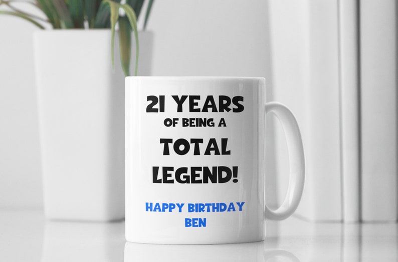 K&ouml;nnte beinhalten: Wei&szlig;e Keramiktasse mit schwarzem Text, der "21 Jahre, ein echter Held! Alles Gute zum Geburtstag Ben" lautet.