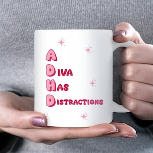 May include: White ceramic mug with the text "ADIVA HAS DISTRACTIONS" in pink letters. Pink starburst graphics are scattered around the text. The mug is held by a person with pink glitter nail polish.