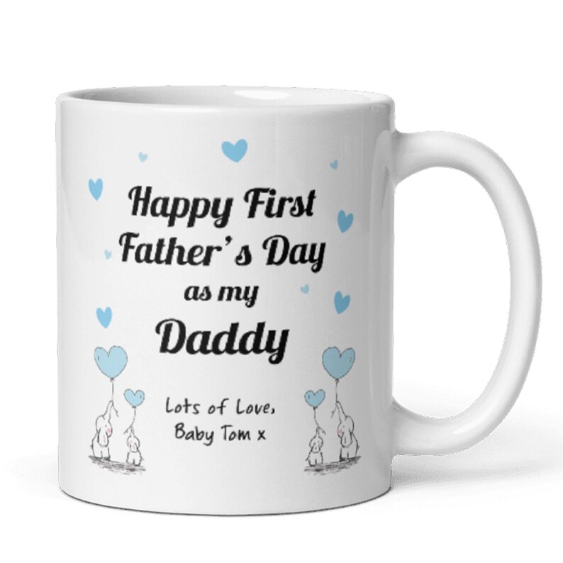 Puede incluir: Taza de cer&aacute;mica blanca con un dise&ntilde;o azul y negro. La taza dice "Happy First Father's Day as my Daddy" con dos corazones azules y dos elefantes sosteniendo globos azules. El texto "Lots of Love, Baby Tom x" est&aacute; en la parte inferior.