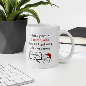 Op de afbeelding: Witte keramische mok met een humoristisch kerstthema. De mok heeft de tekst "I took part in Secret Santa and all I got was this lousy mug." Een cartoon kerstman met een tekstballon met "Ho F**king Ho" staat er ook op.