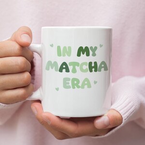 Może przedstawiać: Biały kubek ceramiczny z zielono-białym wzorem, na kt&oacute;rym widnieje napis "In My Matcha Era".