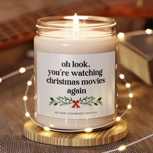 Könnte beinhalten: Eine brennende Sojawachskerze in einem durchsichtigen Glas mit dem Text "oh look, you're watching christmas movies again". Die Kerze steht auf einem runden Holzuntersetzer, mit einer Lichterkette und einer Gitarre im Hintergrund.