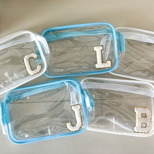 Könnte beinhalten: Fünf transparente Plastik-Reißverschlusstaschen mit blauem Besatz. Jede Tasche hat einen weißen Filzbuchstaben auf der Vorderseite, der das Wort "CLJS" buchstabiert.