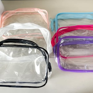 May include: A collection of clear, transparent cosmetic bags with colored trim and handles. The bags are in various colors, including pink, blue, purple, and black. The bags are designed to hold makeup and other beauty products.