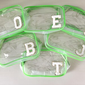 May include: Clear, rectangular cosmetic bags with neon green trim. Each bag has a white, embroidered letter: D, E, S, B, J, and T. The bags are arranged on a white surface.