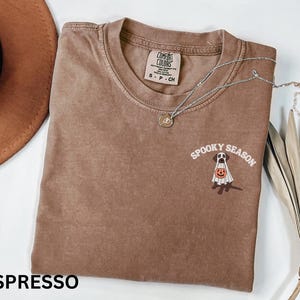 Może przedstawiać: Koszulka z kr&oacute;tkim rękawem w kolorze espresso z okrągłym dekoltem. Na piersi znajduje się grafika przedstawiająca psa w białym prześcieradle ducha, trzymającego pomarańczową dynię, z białym napisem SPOOKY SEASON wygiętym w łuk powyżej.