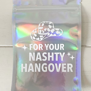 以下が含まれることがあります： 白いカウボーイハットのグラフィックと「FOR YOUR NASHTY HANGOVER」のテキストが入った、ホログラフィックシルバーのポーチ。ポーチはジッパー開閉式で、光沢のある虹色の仕上がりです。