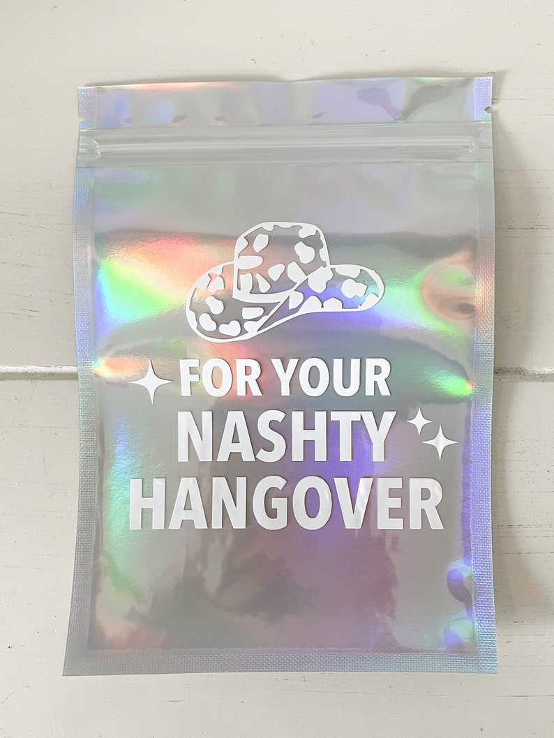 以下が含まれることがあります： 白いカウボーイハットのグラフィックと「FOR YOUR NASHTY HANGOVER」のテキストが入った、ホログラフィックシルバーのポーチ。ポーチはジッパー開閉式で、光沢のある虹色の仕上がりです。