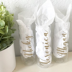 Könnte beinhalten: Drei klare Champagnerflöten aus Glas mit goldener Beschriftung. Die Flöten sind in weißes Seidenpapier eingewickelt. Die Namen "Nicky", "Veronica" und "Megan" sind auf die Flöten geschrieben.