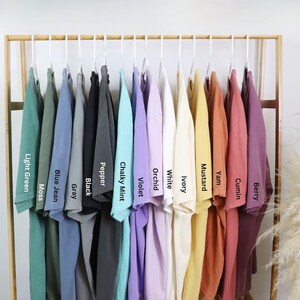Może przedstawiać: Kolekcja koszulek z kr&oacute;tkim rękawem zawieszonych na drewnianym stojaku, prezentowana w spektrum kolor&oacute;w, w tym Light Green, Moss, Blue Jean, Gray, Black, Pepper, Chalky Mint, Violet, Orchid, White, Ivory, Mustard, Yam, Cumin oraz Berry.