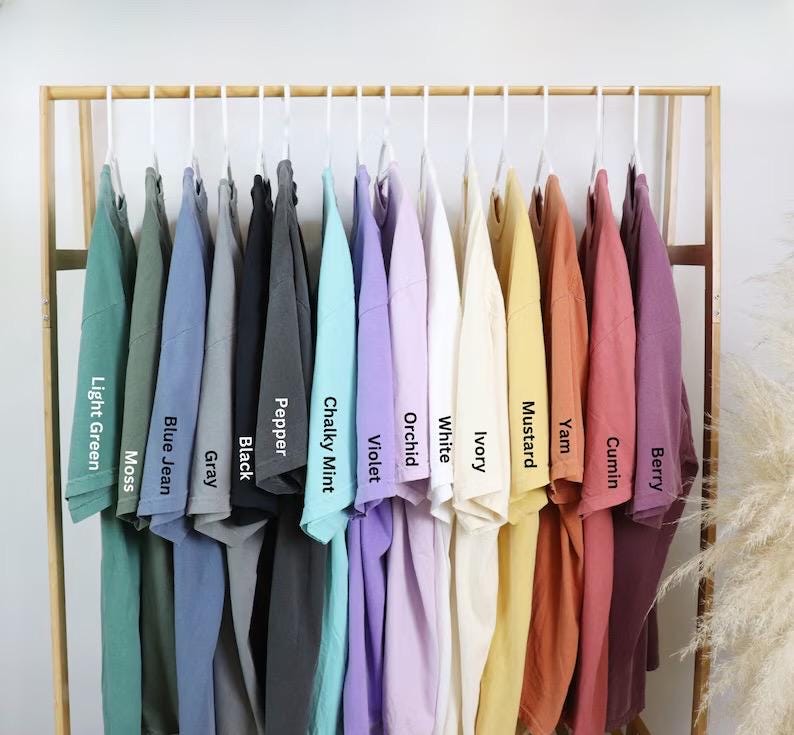Może przedstawiać: Kolekcja koszulek z kr&oacute;tkim rękawem zawieszonych na drewnianym stojaku, prezentowana w spektrum kolor&oacute;w, w tym Light Green, Moss, Blue Jean, Gray, Black, Pepper, Chalky Mint, Violet, Orchid, White, Ivory, Mustard, Yam, Cumin oraz Berry.
