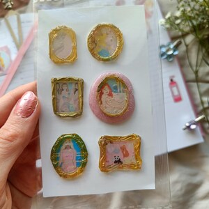以下が含まれることがあります： 透明なプラスチック製のスリーブに入った、6つのミニチュア額入りアート作品のセット。 金色のフレームには、パステルカラーの漫画のキャラクターが描かれています。 フレームは楕円形、長方形、正方形です。