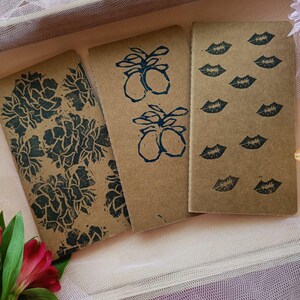 Peut inclure: Trois carnets marrons avec des motifs différents. Le premier carnet a un motif floral, le deuxième a une image répétée d'un fruit avec des feuilles, et le troisième a une image répétée d'une empreinte de lèvres.