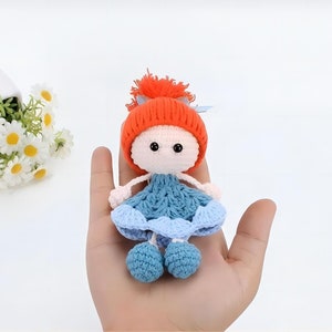 Könnte beinhalten: Eine gehäkelte Puppe mit roter Mütze, blauem Kleid und orangefarbenen Haaren. Die Puppe hält eine blaue Blume in der Hand.