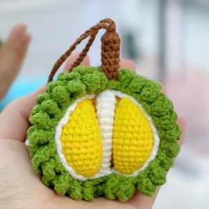 以下が含まれることがあります： 緑色のトゲのある外側と黄色の内側を持つ、かぎ針編みで作ったドリアンフルーツのキーホルダー。キーホルダーは手に持たれています。