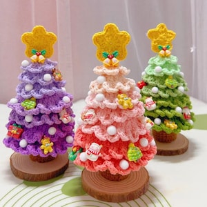 Puede incluir: Tres árboles de Navidad de crochet en morado, rosa y verde con pompones blancos y varios adornos. Cada árbol está sobre una base de madera.