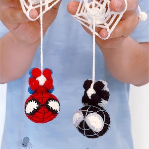 May include: Two crocheted figures hanging from white yarn strings attached to white crocheted web squares. One figure is red and blue with large white eyes, and the other is black and white with large white eyes, both featuring round heads.