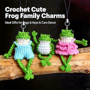 May include: Crochet frog family charms in green, white, and pink. One frog wears blue overalls, another has a white belly, and the third wears a pink dress. The charms hang from silver keychains and are displayed on a wooden branch.