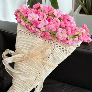 May include: A handmade crochet bouquet featuring pink and green flowers spilling from a cream-colored cone-shaped holder. The bouquet is tied with a sheer, light-tan ribbon. The flowers are densely packed, creating a full, textured appearance.