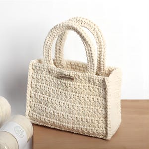 May include: Cream-colored, handmade crochet tote bag with two rounded handles. The bag features a textured stitch pattern and a small metal tag that reads "handmade". The bag is sitting on a wooden surface, with two spools of yarn visible in the background.