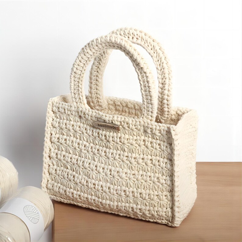 May include: Cream-colored, handmade crochet tote bag with two rounded handles. The bag features a textured stitch pattern and a small metal tag that reads "handmade". The bag is sitting on a wooden surface, with two spools of yarn visible in the background.